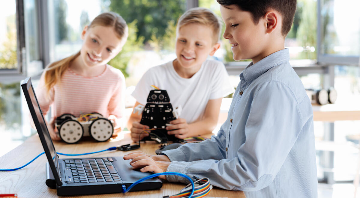 Una guida per accompagnare i bambini nella programmazione - Interesting task. Joyful little boy using the laptop and changing settings of his own robot while his friends observing the process with curiosity