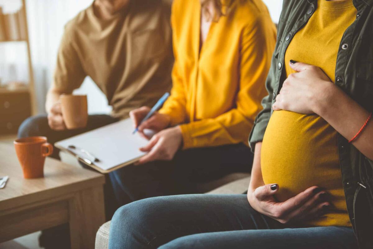 Maternità surrogata: cos'è, come funziona in Italia - Focus on pregnant woman touching her abdomen with gentleness. Married couple is putting signature on document while sitting on background. Surrogate motherhood concept