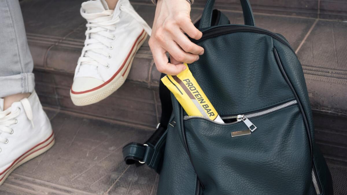 Quali sono le migliori barrette proteiche? - Close-up of a girl crouched on the steps for a light snack. She pulls a protein bar out of her women's backpack. Diet and monitor nutrition