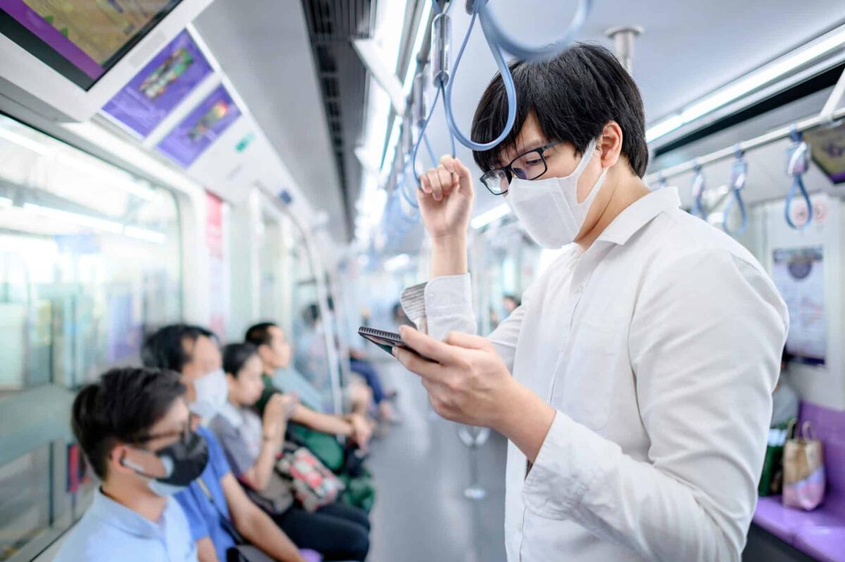 Preoccupa in Cina la variante XBB del Covid - Asian man wearing surgical face mask using smartphone on skytrain or urban train. Wuhan coronavirus (COVID-19) outbreak prevention in public transportation. Health awareness for pandemic protection