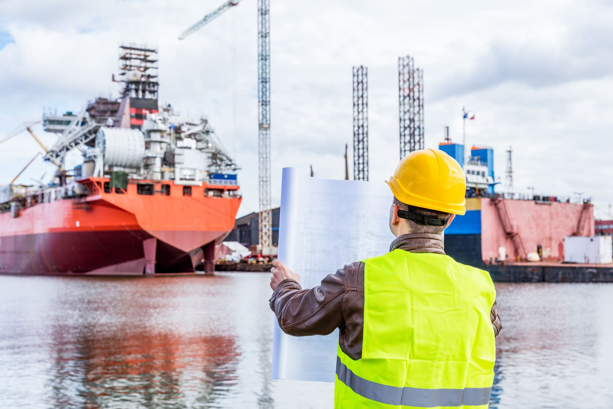 Shipbuilding engineer checking documents and plans of construction at the dockside in a port. Wearing safety helmet and yellow vest, holding folded papers.
