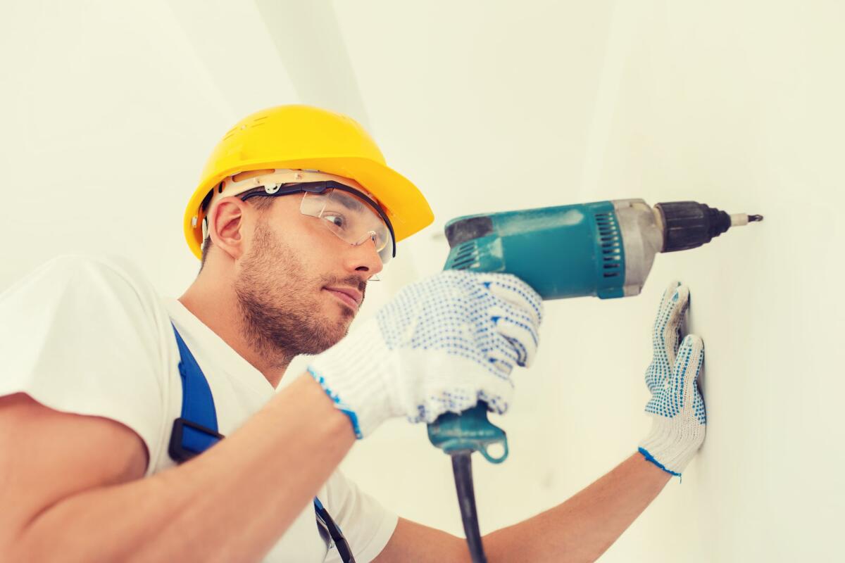 Scegliere l’attrezzatura da lavoro: una guida per coloro che vogliono investire nel futuro della loro professione ed azienda - building, working equipment and people concept - builder in hardhat perforating wall with drill indoors