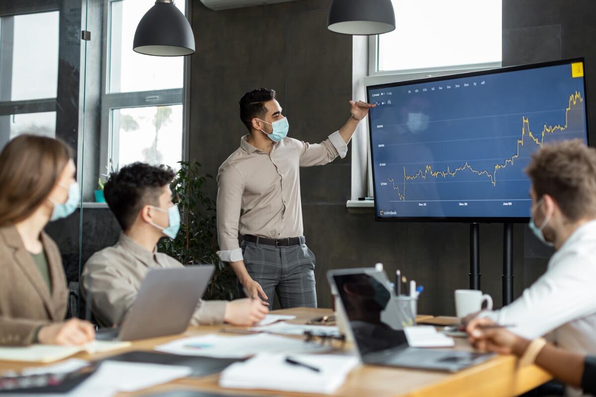 Nuovo piano pandemico in Italia: tra vaccini Novavax e controllo dei contagi - Modern Corporate Meeting Concept. Young Businessman In Medical Face Mask Giving Speech During Seminar With Coworkers In Office, Showing Growth Chart On Screen Monitor Standing In Board Room