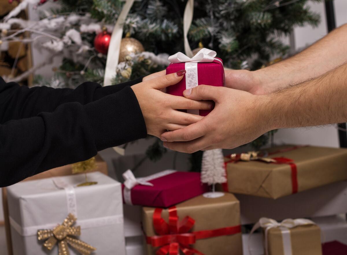 Idee regalo per Natale da acquistare durante il Black Friday - Hands of man giving a Christmas gift to girlfriend. Decorated christmas tree on background