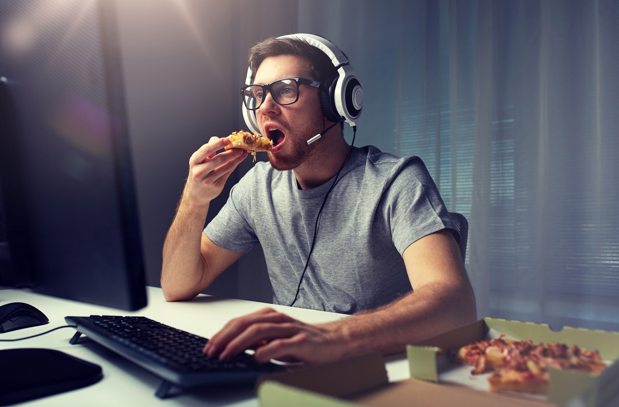 technology, gaming, entertainment, let's play and people concept - young man in headset with pc computer eating pizza while playing game at home and streaming playthrough or walkthrough video