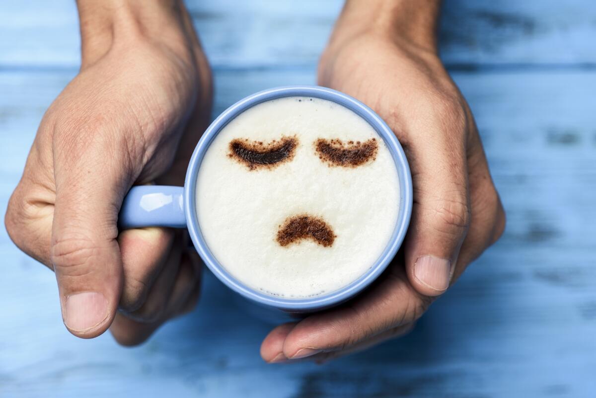 Blue Monday: oggi è il giorno più triste dell'anno (o forse no) - high-angle shot of a young caucasian man with a blue cup of cappuccino with a sad face drawn with cocoa powder on the milk foam, on a blue rustic table