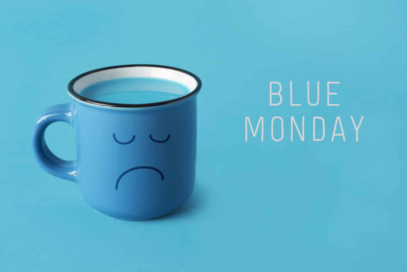 closeup of a blue mug, with a sad face drawn in it, and the text blue monday on a blue background