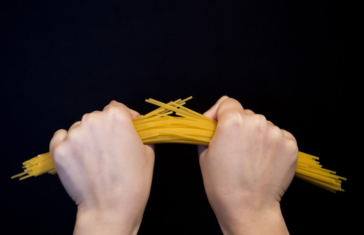 Perché quando spezziamo gli spaghetti si dividono in più pezzi? - horizontal image of female hands clenching in fists break in half a bunch of raw long yellow thin spaghetti pasta on a black background